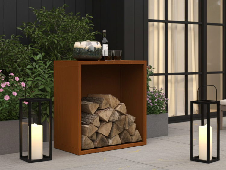 Brændeskjul i cortenstål med stablet brænde, placeret på moderne terrasse ved sort facade med lanterner i aftenlys.