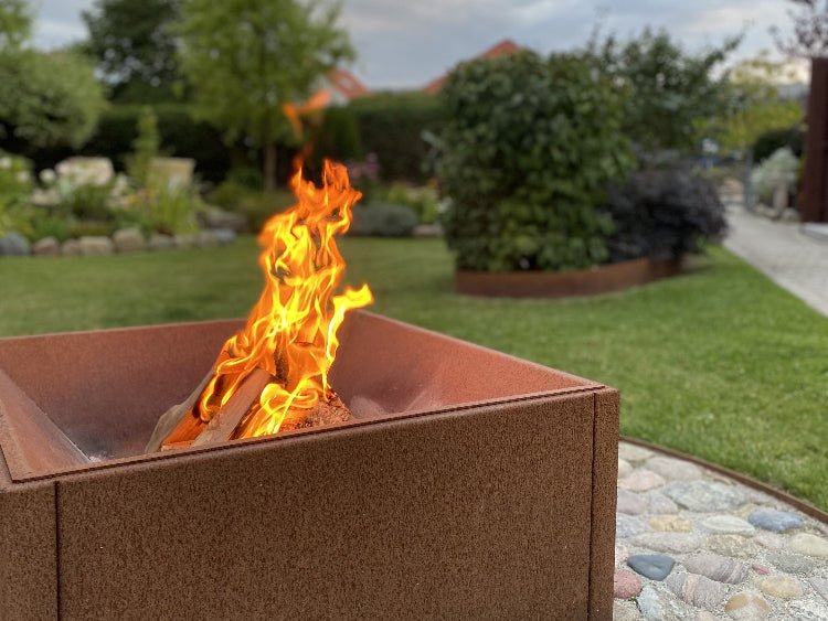 Firkantet bålkasse i cortenstål med levende ild placeret i grøn have med græs og bede