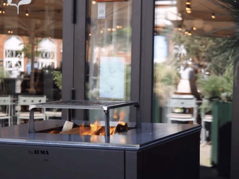 CUBY bålgrill i sortlakeret stål i udendørs restaurantmiljø med glasfacade og levende ild