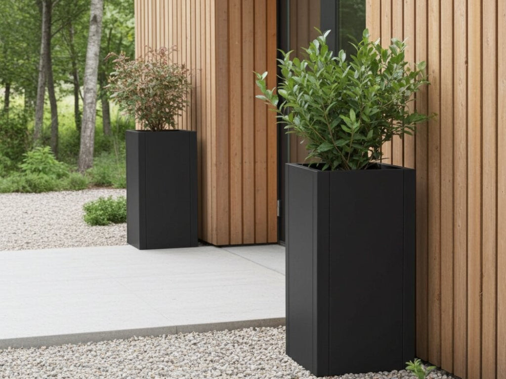 Høje sorte plantekrukker i stål med grønne buske ved husets indgang