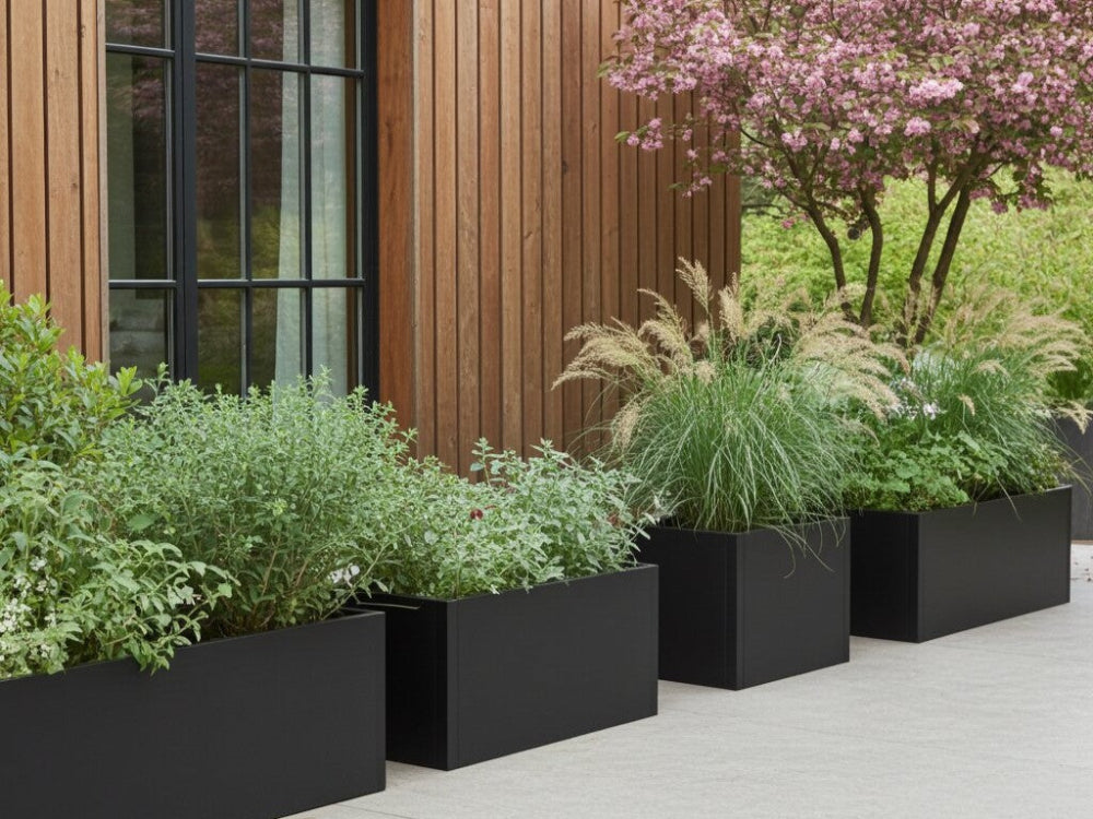 Sorte plantekasser i stål med stauder og prydgræsser placeret i række langs træfacade