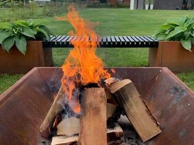 Bålkar i cortenstål med brændestykker og levende flammer, placeret i have med integrerede bænke og plantekasser