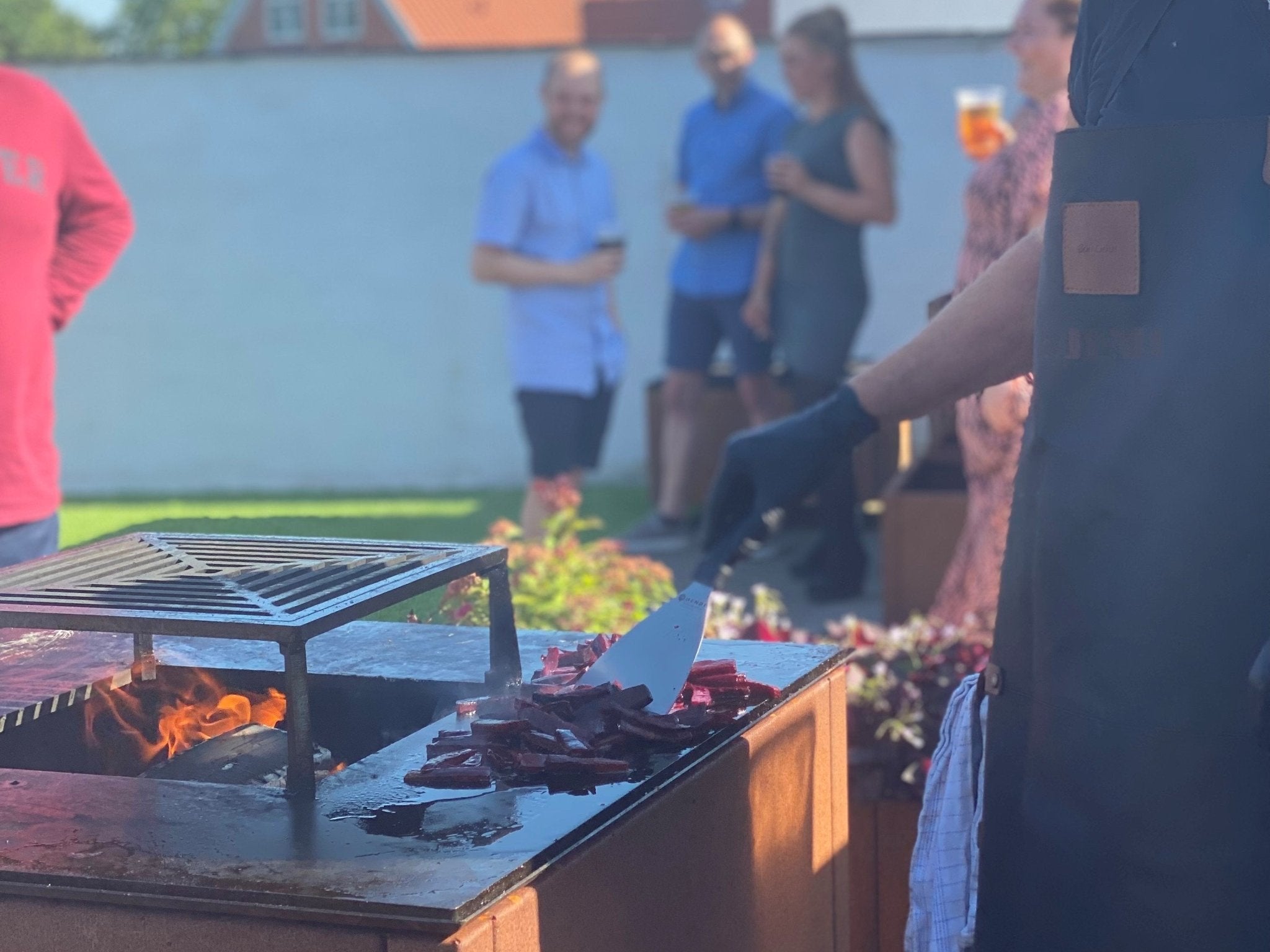 byJEMA bålgrill i cortenstål med sort topplade og grillrist – tilberedning over åben ild i hyggeligt havemiljø med gæster i baggrunden