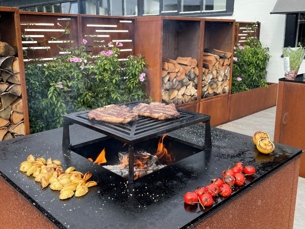 byJEMA bålgrill i cortenstål med sort stål topplade og grillrist – bøffer og grøntsager grilles over åben ild i moderne udekøkkenmiljø