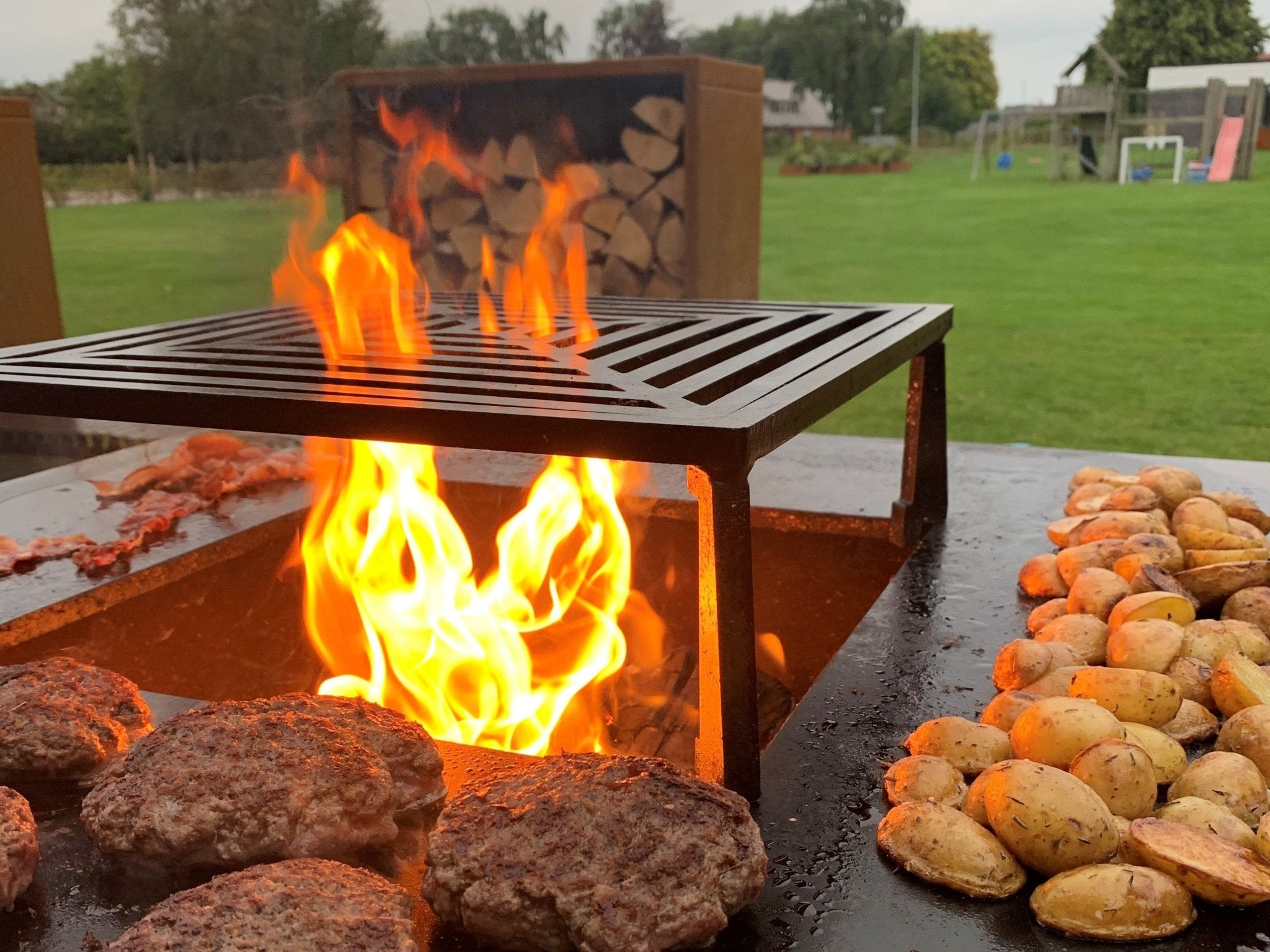 byJEMA bålgrill i cortenstål med sort grillrist og kraftige flammer – bøffer og kartofler tilberedes over åben ild på terrasse i haven