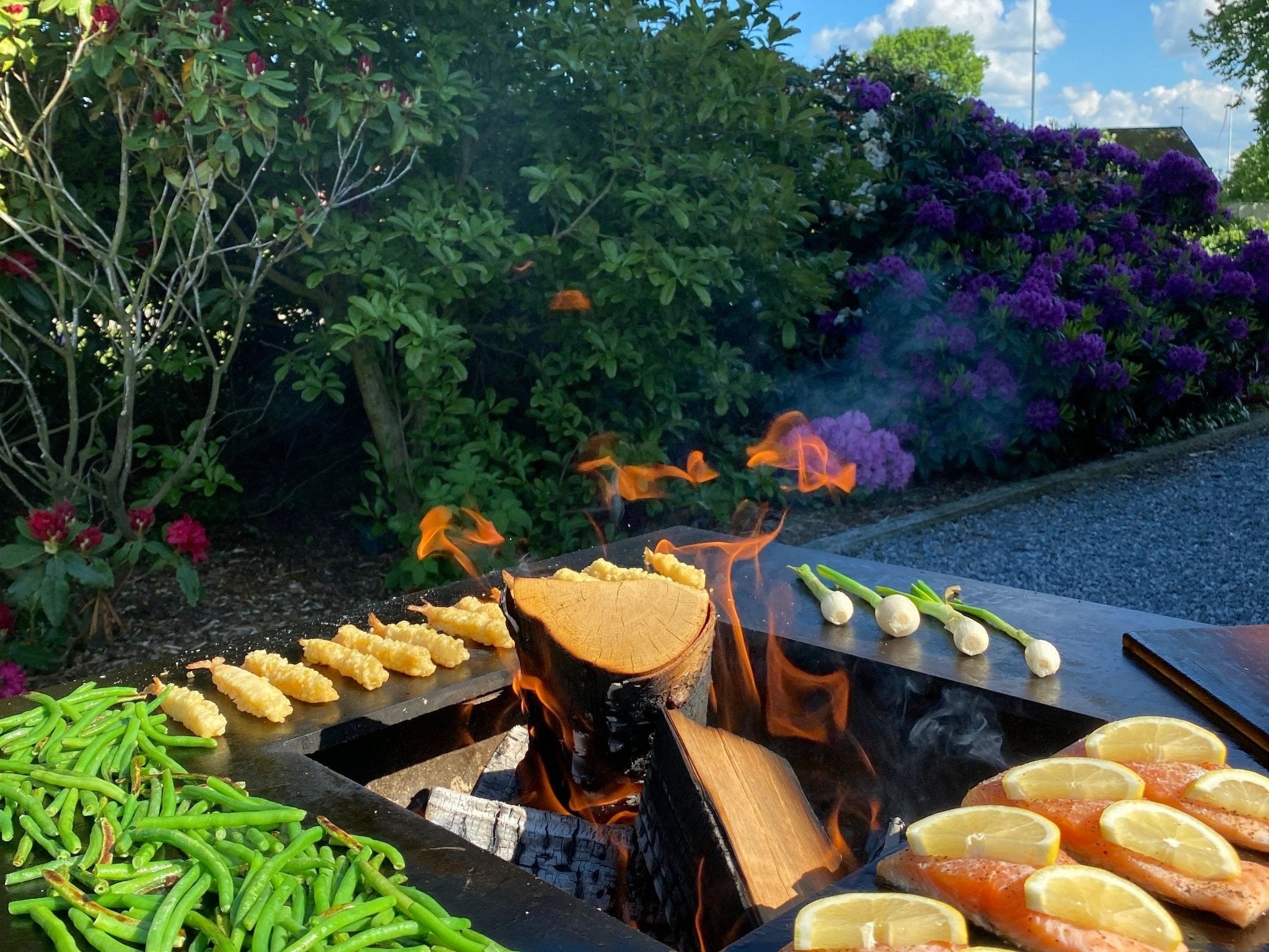 byJEMA bålgrill i cortenstål med sort stål topplade – laks med citron og friske grøntsager tilberedes over åben ild i frodig have