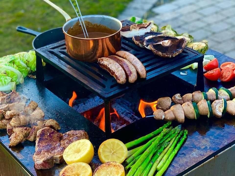 byJEMA bålgrill i cortenstål med sort grillrist – pande, pølser, grøntsager og kød tilberedes over åben ild på udendørs stålgrill