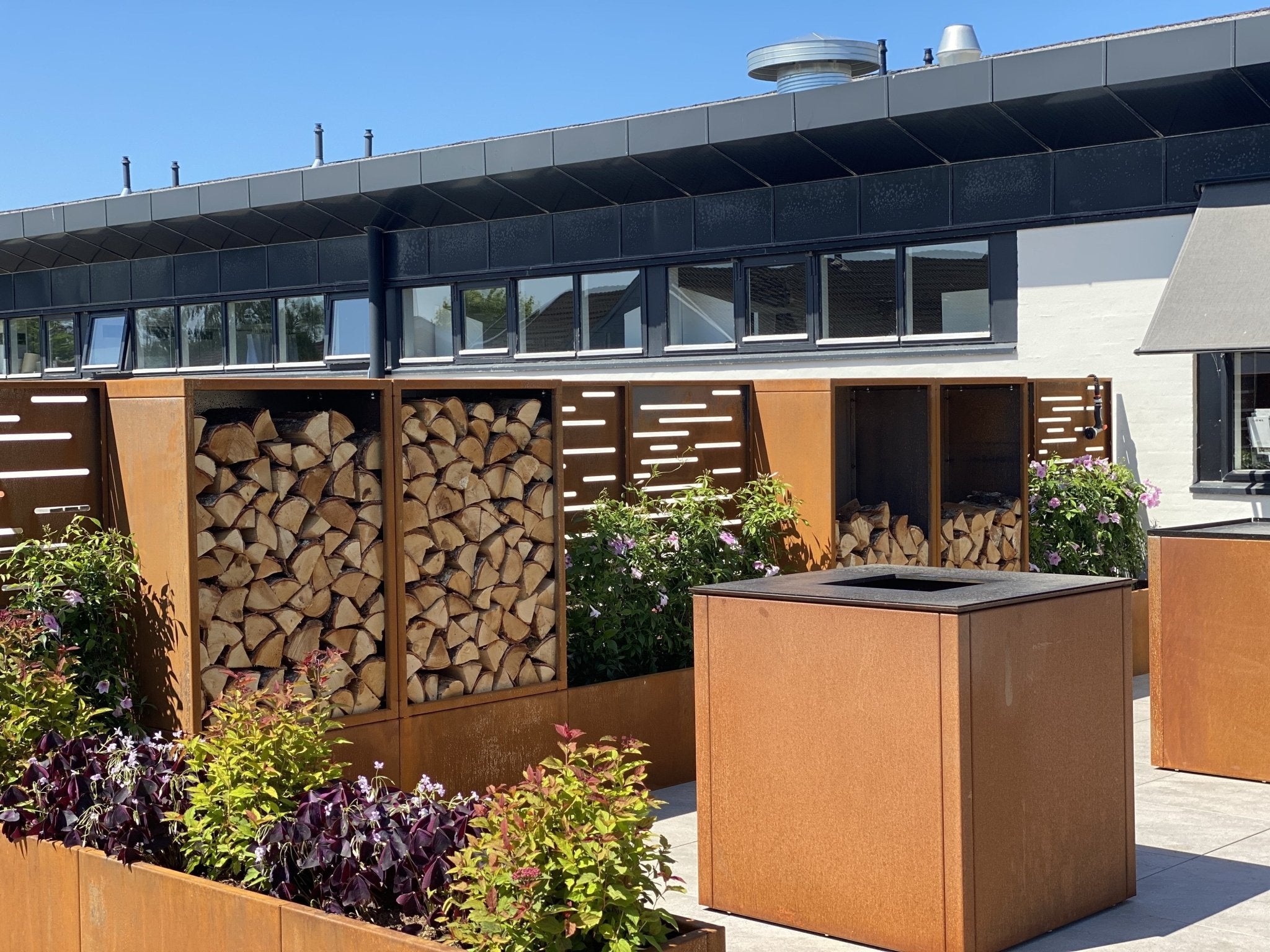 byJEMA bålgrill i cortenstål på moderne terrasse med integreret vedopbevaring og matchende plantekasser – stilrent udendørs ildsted i arkitektonisk havemiljø