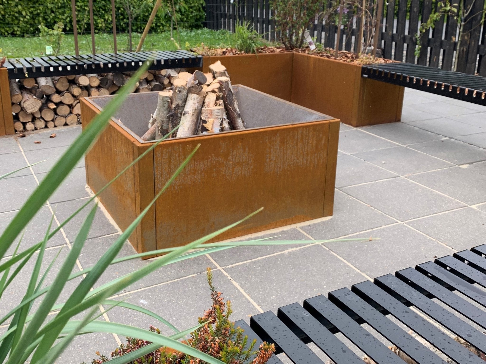 Firkantet bålkar i cortenstål med brænde, omgivet af integrerede siddebænke og brændestabel på fliseterrasse