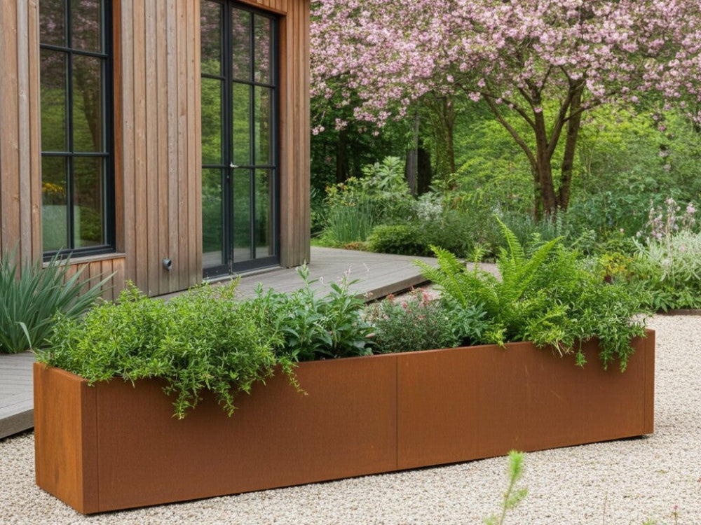 Lang CUBY plantekasse i cortenstål med stauder og bregner foran træbeklædt hus