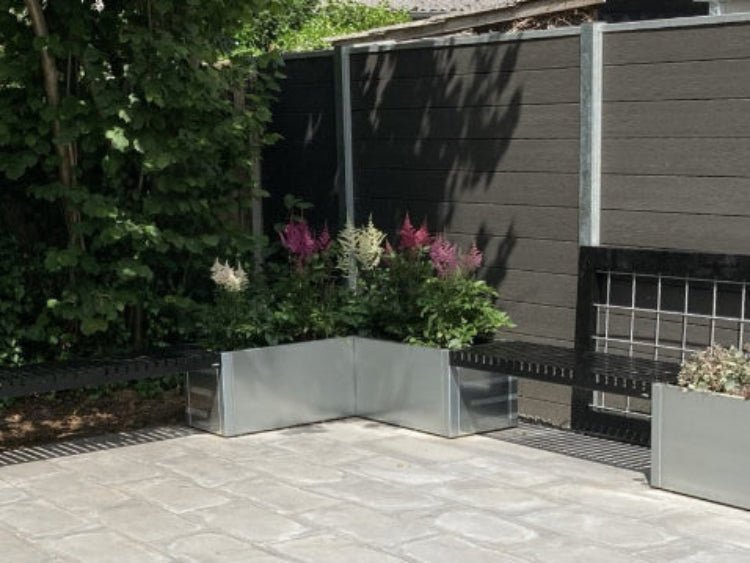 Vinkel plantekasse i galvaniseret stål med blomstrende planter langs hegn