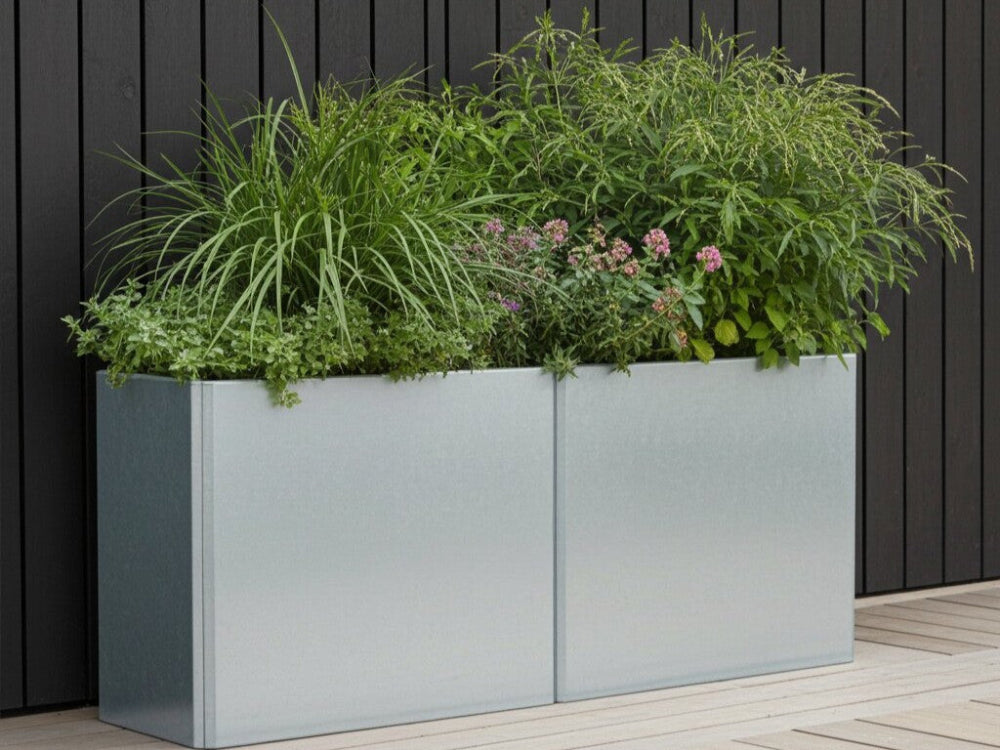 Lang EDGY plantekasse i galvaniseret stål med tæt grøn beplantning foran mørk facade