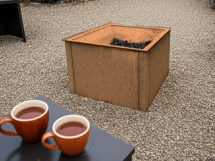 Firkantet bålkar i cortenstål placeret på grus med gløder i bunden og kaffekopper i forgrunden