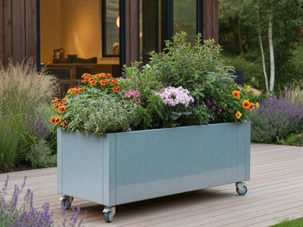 Galvaniseret plantekasse på hjul fyldt med blomster og grønne planter på træterrasse foran moderne bolig.