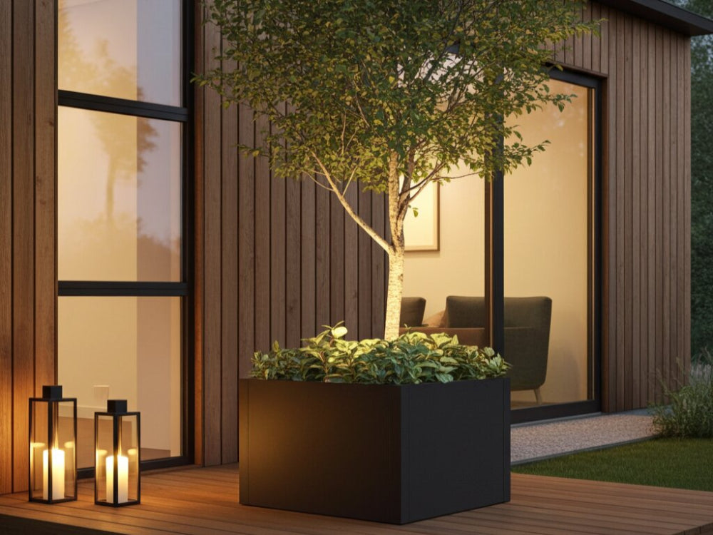 Stor sort plantekasse i stål med lille træ og bundbeplantning på træterrasse i aftenlys