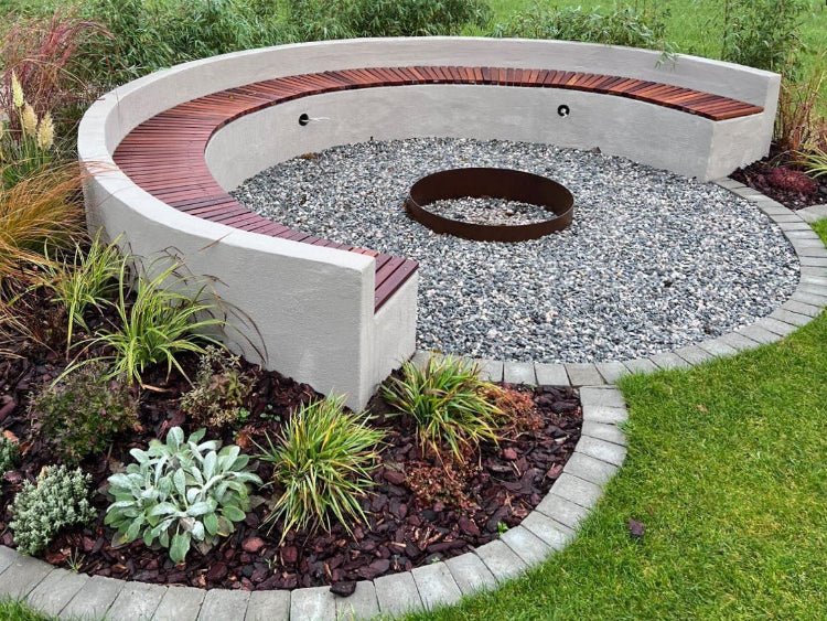 Rund bålplads med bålkar i cortenstål omgivet af buet siddebænk og grusbelægning i haven