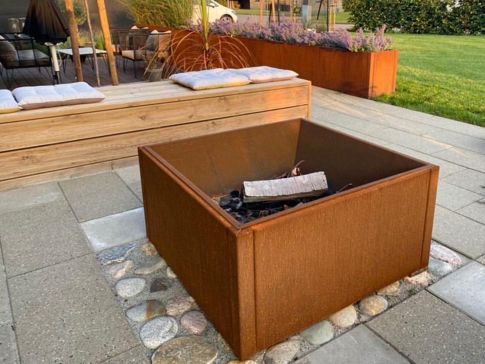 Firkantet bålkasse i cortenstål placeret på terrasse uden ild, integreret i moderne havemiljø med plantekasser og træbænk