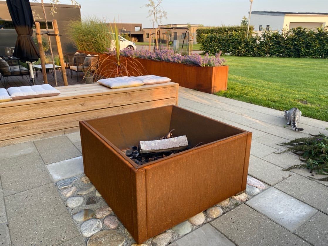 Firkantet bålkar i cortenstål placeret på fliseterrasse med træbænk og cortenstål plantekasser i baggrunden