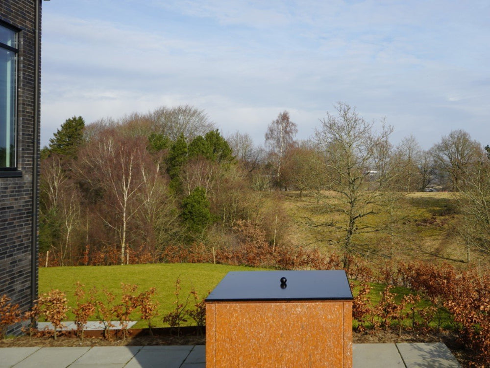 Låg til bålgrill i sort stål placeret i have med åben udsigt